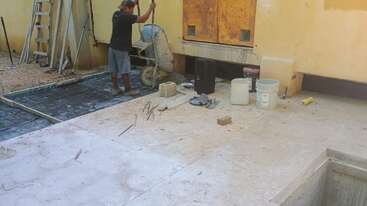 Un trabajador de la construcción empuja una carretilla cerca de un edificio amarillo. Diversos materiales de construcción, herramientas, cubos y ladrillos están esparcidos por el suelo inacabado.