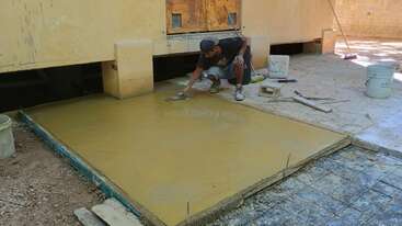 Un hombre alisa hormigón recién vertido con una paleta, trabajando en un patio o una pasarela junto a un edificio. Le rodean herramientas de construcción y cubos.