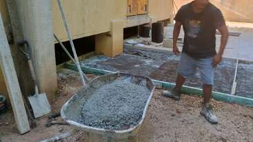 Un trabajador de la construcción junto a una carretilla llena de cemento húmedo. Cerca, las palas descansan contra una pared, y el hormigón se vierte sobre el suelo preparado.