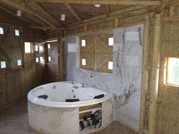 Este baño rústico cuenta con una bañera de hidromasaje redonda con paredes de mármol, rodeada de paredes y techo de bambú, con pequeñas ventanas que proporcionan luz natural.