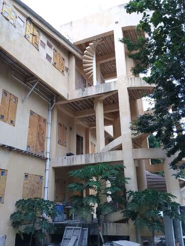 Esta imagen muestra un edificio inacabado o abandonado con ventanas tapiadas, una prominente escalera de caracol, algunas plantas verdes debajo y materiales de construcción esparcidos por el suelo.
