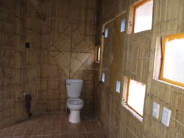 Se trata de un cuarto de baño rústico con paredes de bambú, un inodoro blanco, pequeñas ventanas cuadradas, suelo de baldosas, motivos geométricos en las paredes y un diseño sencillo, natural y terroso.