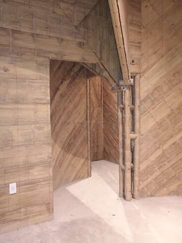 Esta imagen muestra una habitación con paredes y techos de paneles y postes de bambú, con una puerta esquinera y un suelo de hormigón con una toma de corriente.