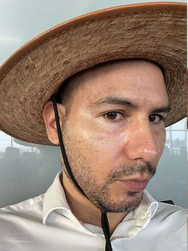 Un hombre con sombrero de paja de ala ancha y camisa blanca de cuello mira a la cámara con expresión seria, visible una ligera barba incipiente y corta.