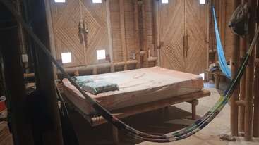 Esta acogedora habitación tiene paredes y puertas de bambú, una cama sencilla con una manta, una hamaca, ventanas pequeñas y una decoración minimalista, que crean un ambiente rústico.