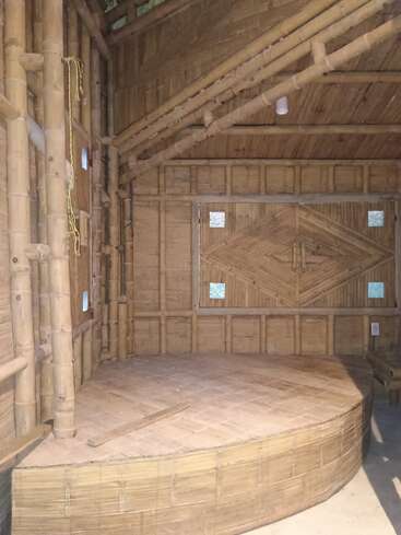 Esta imagen muestra una estructura interior hecha enteramente de bambú, con una plataforma circular elevada, paneles de pared con motivos geométricos y pequeñas ventanas cuadradas que dejan pasar la luz.