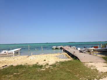 L'image représente une scène de plage sereine avec un quai en bois s'avançant dans l'eau, avec plusieurs bateaux amarrés à proximité, le tout dans un ciel bleu clair.