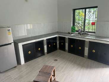 Cette cuisine comprend un réfrigérateur argenté, des armoires noires avec des poignées dorées, un évier en acier inoxydable, des murs carrelés, une grande fenêtre et un petit tabouret en bois.