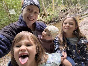 Une femme et trois enfants prennent un selfie dans une forêt, près d'un ruisseau. L'un des enfants tire la langue ; tout le monde a l'air joyeux et enjoué.