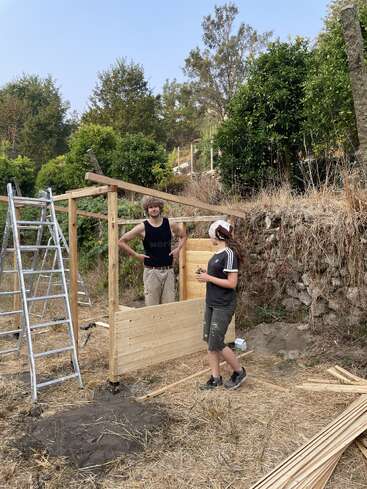Two people are building a wooden structure outdoors, surrounded by trees and dry grass. A ladder stands nearby, with scattered planks and tools on the ground.