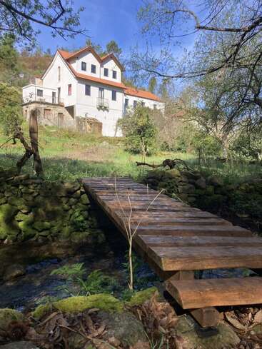 A charming white house with a red roof stands on a grassy hill, behind a rustic wooden footbridge crossing a small mossy stone-lined stream. Peaceful nature.