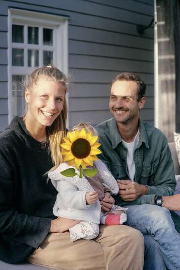 Une femme et un homme souriants sont assis dehors et posent pour une photo. Un enfant est assis sur les genoux de la femme, le visage caché par un grand tournesol.