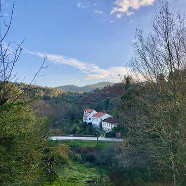 A white house with red roof sits nestled in a lush green valley, surrounded by trees, hills, and a bright, blue sky with scattered clouds.