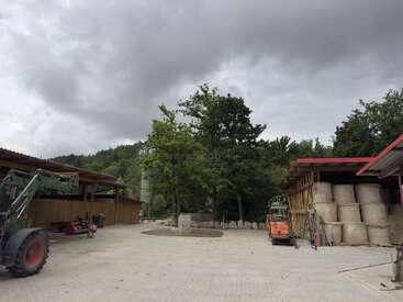 Esta imagen muestra un corral con un tractor, pacas de heno apiladas bajo un refugio, árboles en el centro y un cielo nublado que sugiere una inminente tormenta.
