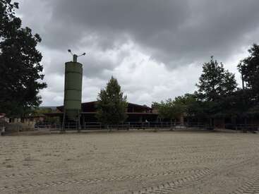 Un cielo nublado se cierne sobre una zona de arena vacía con huellas de neumáticos visibles, rodeada de árboles, un alto silo verde y edificios con forma de granero al fondo.