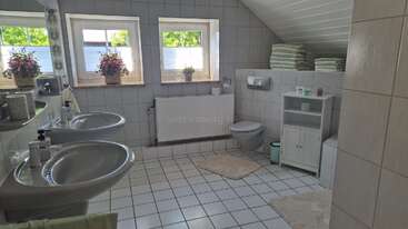Este cuarto de baño es espacioso, luminoso y limpio, con dos lavabos, ventanas, azulejos claros, toallas apiladas, alfombras acogedoras, plantas y adornos florales en el alféizar.