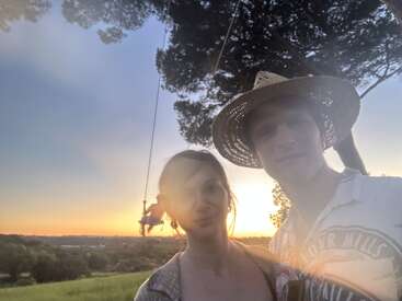 Un couple se tient dehors au coucher du soleil, souriant pour un selfie. La lumière du soleil crée un reflet sur l'objectif. À l'arrière-plan, quelqu'un se balance sur une corde près d'un grand arbre.