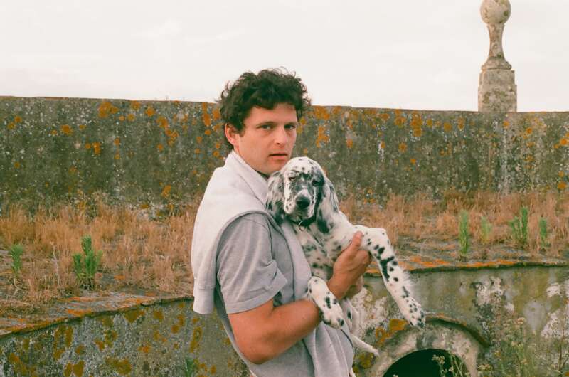 Un jeune homme aux cheveux bouclés, vêtu d'une chemise et d'un pull gris clair, tient dans ses bras un chiot tacheté à l'extérieur, adossé à un mur de pierre moussu, sous un ciel couvert.