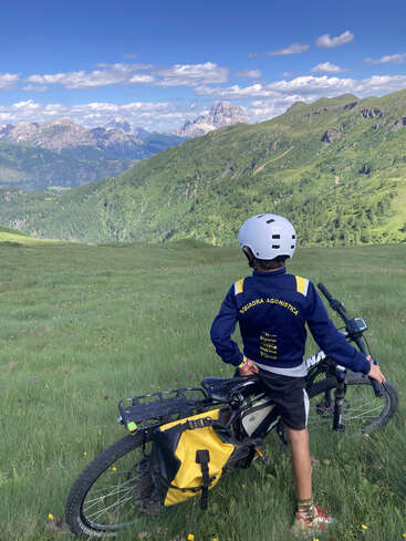 Un joven ciclista, con casco y chaqueta azul, está de pie junto a su bicicleta en una colina cubierta de hierba, admirando el impresionante paisaje montañoso bajo un cielo azul brillante.