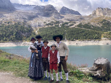 Una familia vestida con ropa tradicional alpina posa junto a un tranquilo lago de montaña, rodeada de picos rocosos, exuberante vegetación y un cielo sereno y nublado.