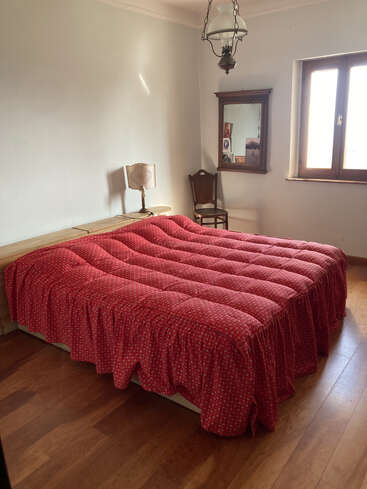 Un acogedor dormitorio con suelos de madera, una gran cama cubierta con un edredón rojo, mesitas auxiliares de madera, una lámpara vintage, un espejo, una silla y luz natural.