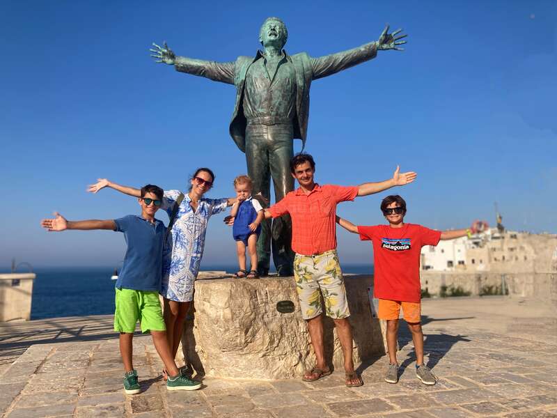 Una familia de cinco miembros posa alegremente frente a una gran estatua con los brazos extendidos, imitando su postura, en un día soleado junto al mar.