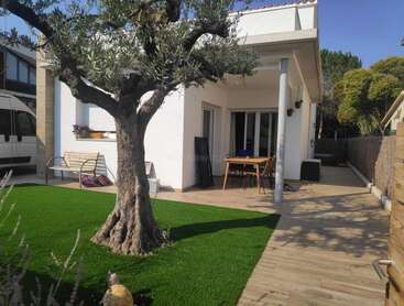 La imagen muestra un sereno patio trasero con una casa blanca, un árbol y un patio con mesa y sillas, rodeado de exuberante vegetación y un cielo azul despejado.