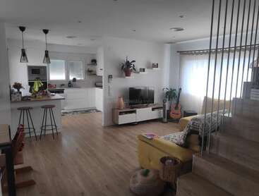 La imagen muestra un moderno espacio diáfano con cocina, salón y escalera, paredes blancas, suelos de madera y una estética minimalista.