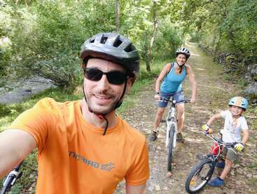 La imagen muestra a una familia de tres miembros en bicicleta de montaña, con casco, posando para un selfie en un camino de tierra en una zona boscosa en un día soleado.