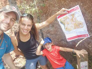 La imagen muestra a una familia de tres miembros posando para una foto frente a un acantilado rocoso, con la madre sosteniendo un mapa.