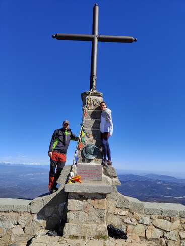 La imagen representa a un hombre y una mujer de pie a ambos lados de un monumento de piedra con una gran cruz encima, con un fondo montañoso y un cielo azul despejado.