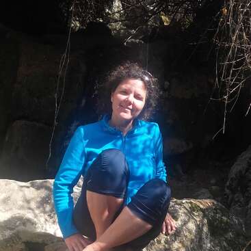 The image depicts a woman sitting on a rock, wearing a blue jacket and dark pants, with a serene expression, surrounded by a natural setting with rocks and vines.