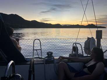 Two people relax on a sailboat at sunset, gazing at calm water and silhouetted hills. The tranquil scene captures a peaceful and serene evening moment.