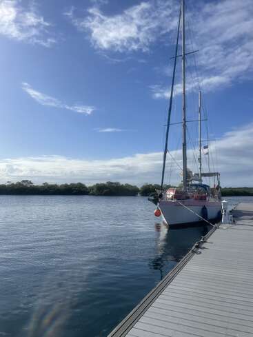 Ein Segelboot liegt an einem Holzsteg auf ruhigem Wasser unter einem teilweise bewölkten blauen Himmel, und in der Ferne sieht man Bäume am Horizont.