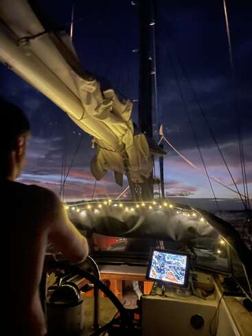 Jemand steuert ein Segelboot in der Abenddämmerung. Der Himmel ist dunkel und das Licht wird schwächer; Lichterketten machen das Cockpit schön und ein Navigationsbildschirm leuchtet hell.