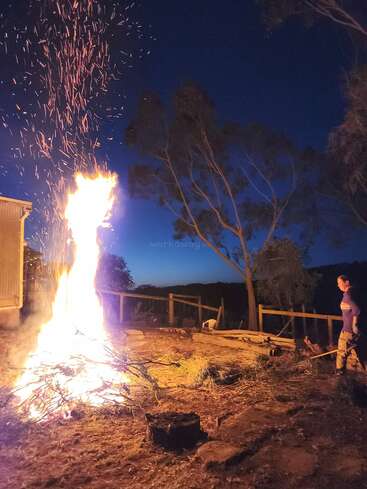 Uma grande fogueira brilha intensamente, lançando faíscas no céu noturno. Uma pessoa fica por perto observando, cercada por árvores e uma cerca de madeira rústica.