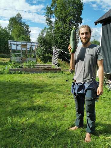 Un homme barbu se tient pieds nus sur l'herbe, tenant une grosse courgette. Derrière lui se trouvent une serre, des plates-bandes, de grands arbres et un ciel bleu.