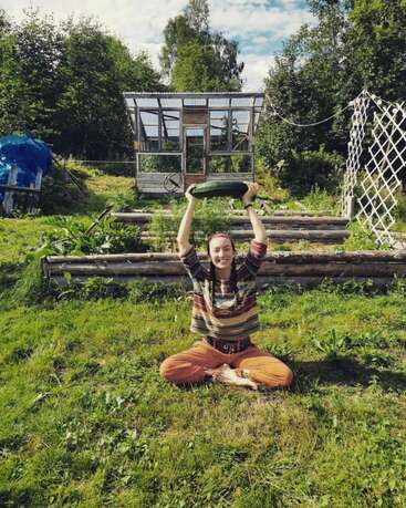 Une femme souriante est assise sur l'herbe, les jambes croisées, tenant une grosse courgette au-dessus de sa tête. Derrière elle, une serre et un jardin luxuriant entourés d'arbres et de plantes vertes.