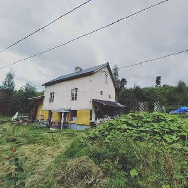 Une maison rustique à deux étages avec des accents jaunes se trouve au milieu d'une verdure luxuriante, de vélos et d'outils de jardinage sous un ciel nuageux, créant une atmosphère de tranquillité et de simplicité rurale.