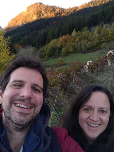 Un hombre y una mujer sonríen mientras se hacen un selfi al aire libre. Detrás de ellos, un frondoso bosque verde y una montaña bañada por el sol crean un hermoso paisaje otoñal natural.