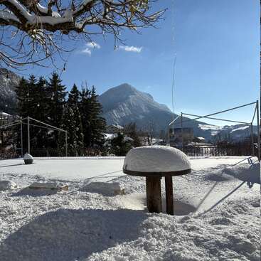 Un paisaje nevado muestra una mesa redonda de madera cubierta de nieve, montañas al fondo, pinos, un cielo azul brillante y abundante luz solar que proyecta sombras.
