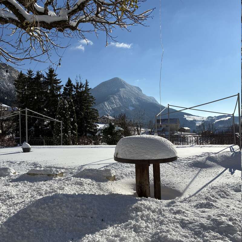 Un paisaje nevado muestra una mesa redonda de madera cubierta de nieve, montañas al fondo, pinos, un cielo azul brillante y abundante luz solar que proyecta sombras.