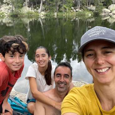 Uma família sorridente de quatro pessoas posa junto a um lago sereno, cercada por árvores e rochas verdejantes, curtindo a natureza e a companhia uns dos outros ao ar livre.