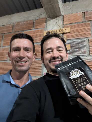 Dois homens sorridentes estão juntos em um ambiente interno, encostados em uma parede de tijolos. Um deles está segurando um saco preto de café "Café La Cima", mostrando-o orgulhosamente para a câmera.