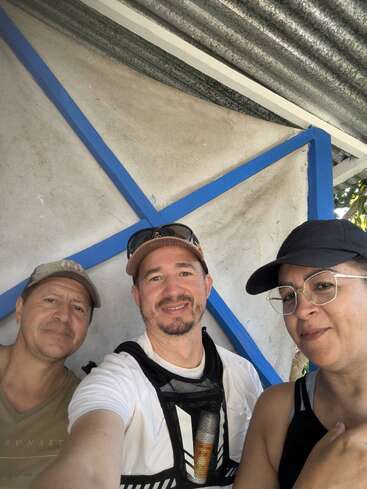 Trois personnes posent pour un selfie, souriant sous un toit ondulé aux poutres bleues. Tous portent des vêtements décontractés et des casquettes, et profitent ensemble d'un moment de détente en plein air.