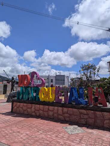 L'enseigne colorée "Yo ❤️ Marquetalia" se trouve sous un ciel bleu vif avec des nuages cotonneux, entourée de plantes, d'une statue et de bâtiments en arrière-plan.