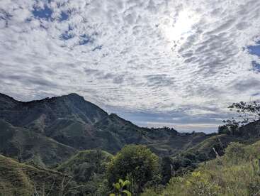 Des collines et des montagnes verdoyantes s'étendent sous un ciel spectaculaire rempli de nuages texturés, la lumière du soleil passant à travers, illuminant le paysage naturel luxuriant et paisible en contrebas.
