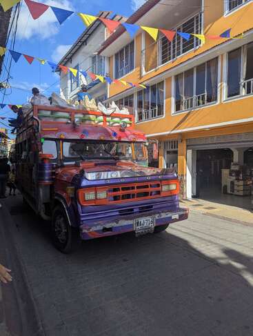 Un bus coloré et décoré roule dans une rue festive bordée de bâtiments lumineux et de drapeaux en guirlande. Des personnes sont assises sur le bus, portant de grands sacs. Scène de jour.