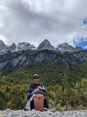 Un hombre sentado en un suelo rocoso con una gran mochila marrón, rodeado de frondosos árboles verdes, con majestuosas montañas nevadas alzándose al fondo.