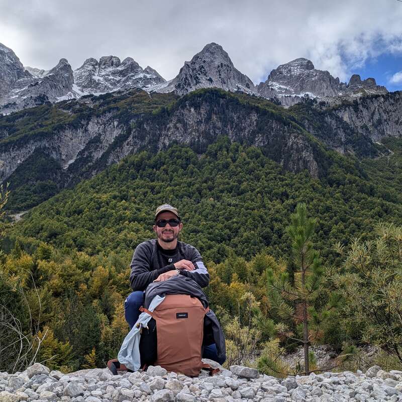 Um homem senta-se em um terreno rochoso com uma grande mochila marrom, cercado por árvores verdes exuberantes, com majestosas montanhas cobertas de neve ao fundo.